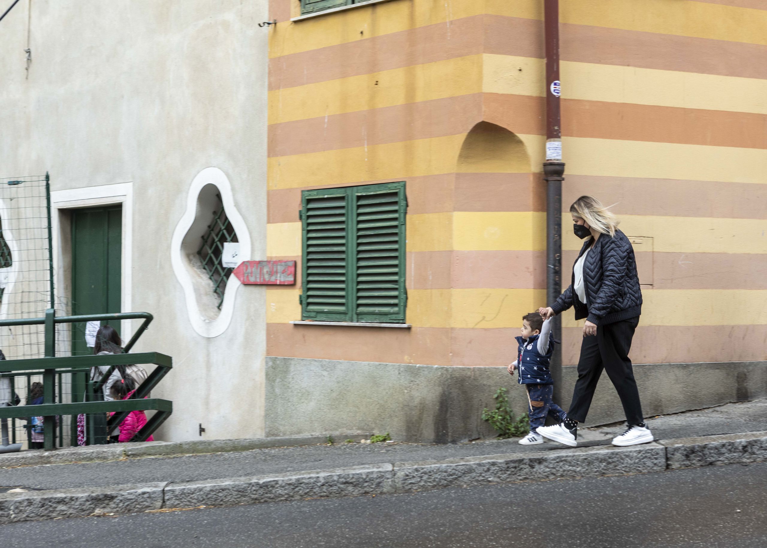 Una donna dà la mano a un bambino mentre camminano sul marciapiede. Sullo sfondo un muro a righe giallo e arancione