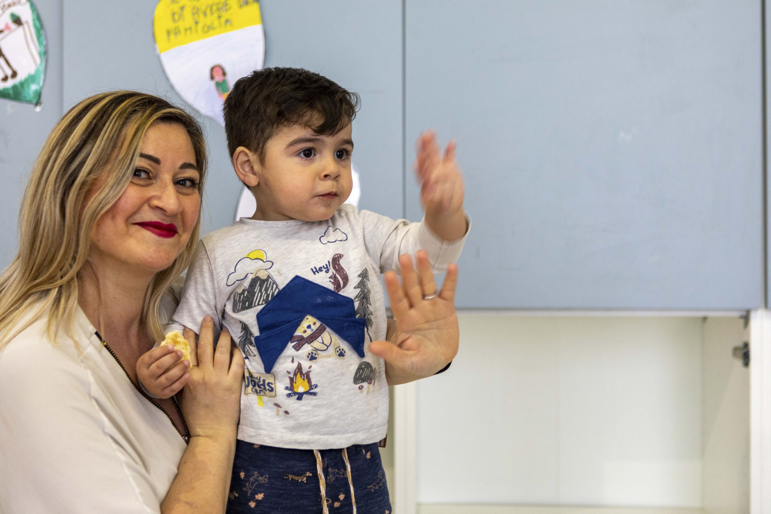 Una donna con i capelli biondi sorride mentre tiene in braccio un bambino piccolo che saluta