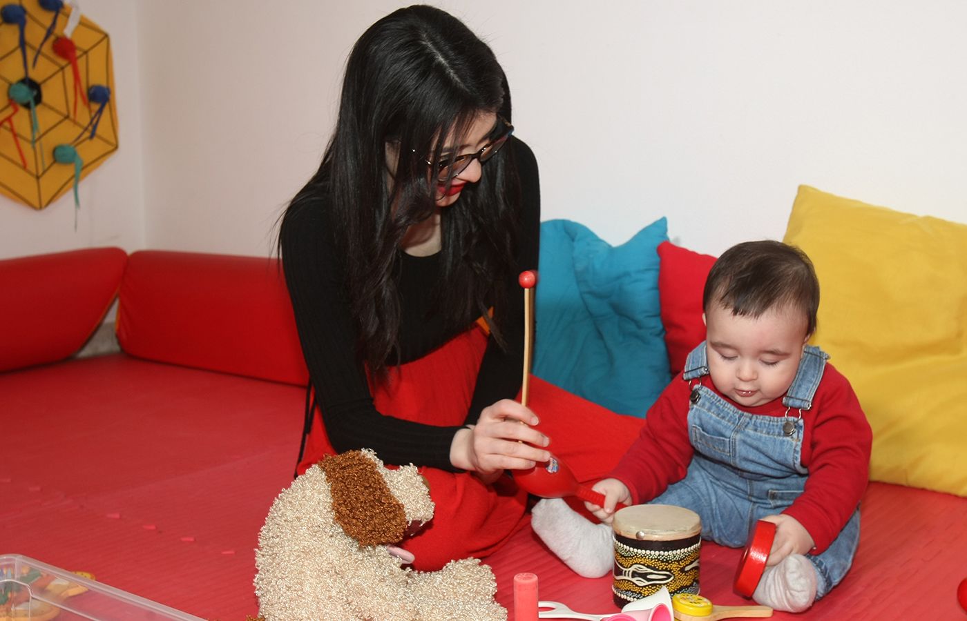 una donna con i capelli neri tiene in mano una bacchetta di uno xilofono mentre gioca con il suo bambino su un tappetino rosso