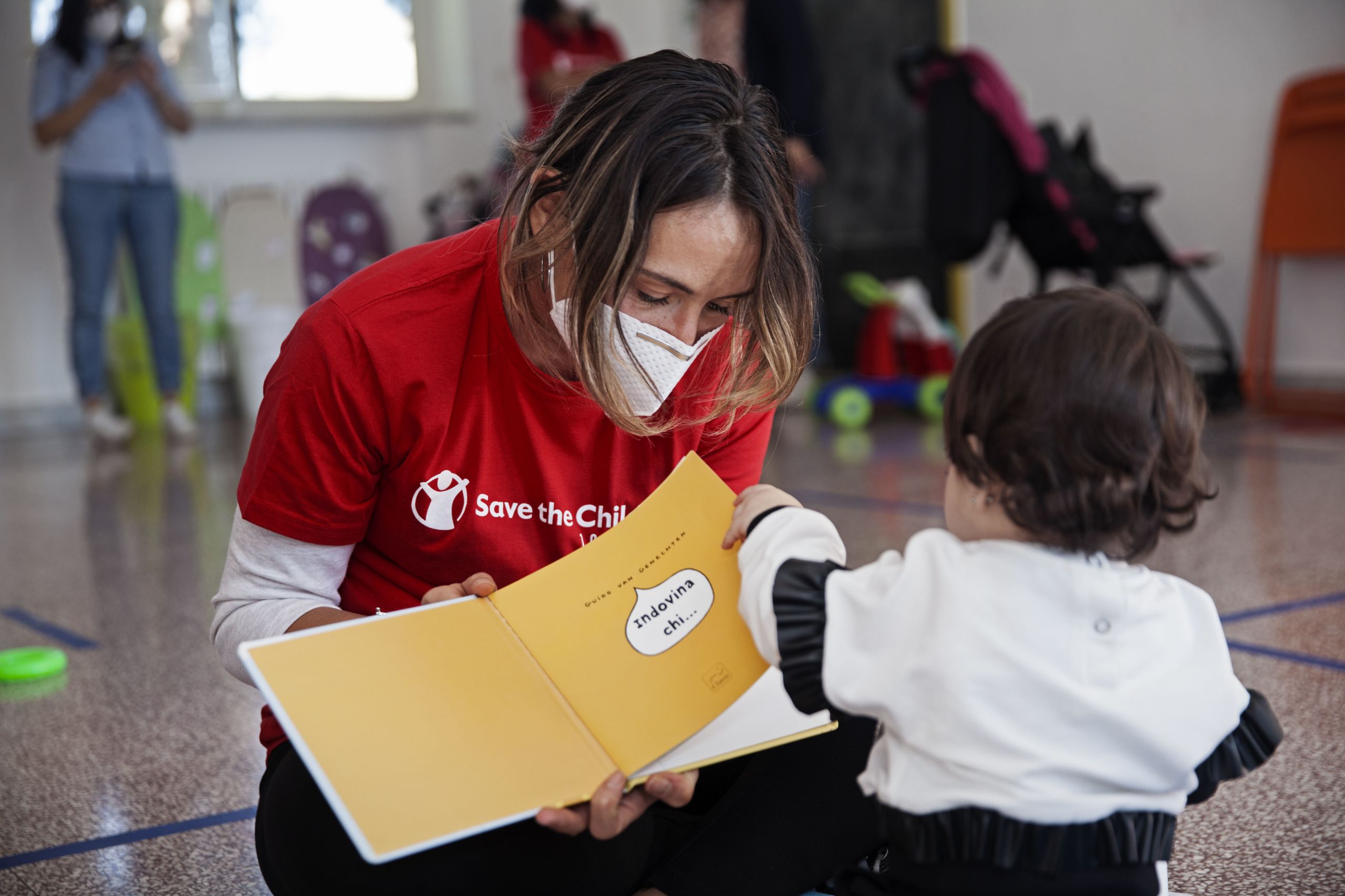 Una operatrice con maglietta rossa e logo Save the Children legge un libro ad una bambina piccola, di spalle